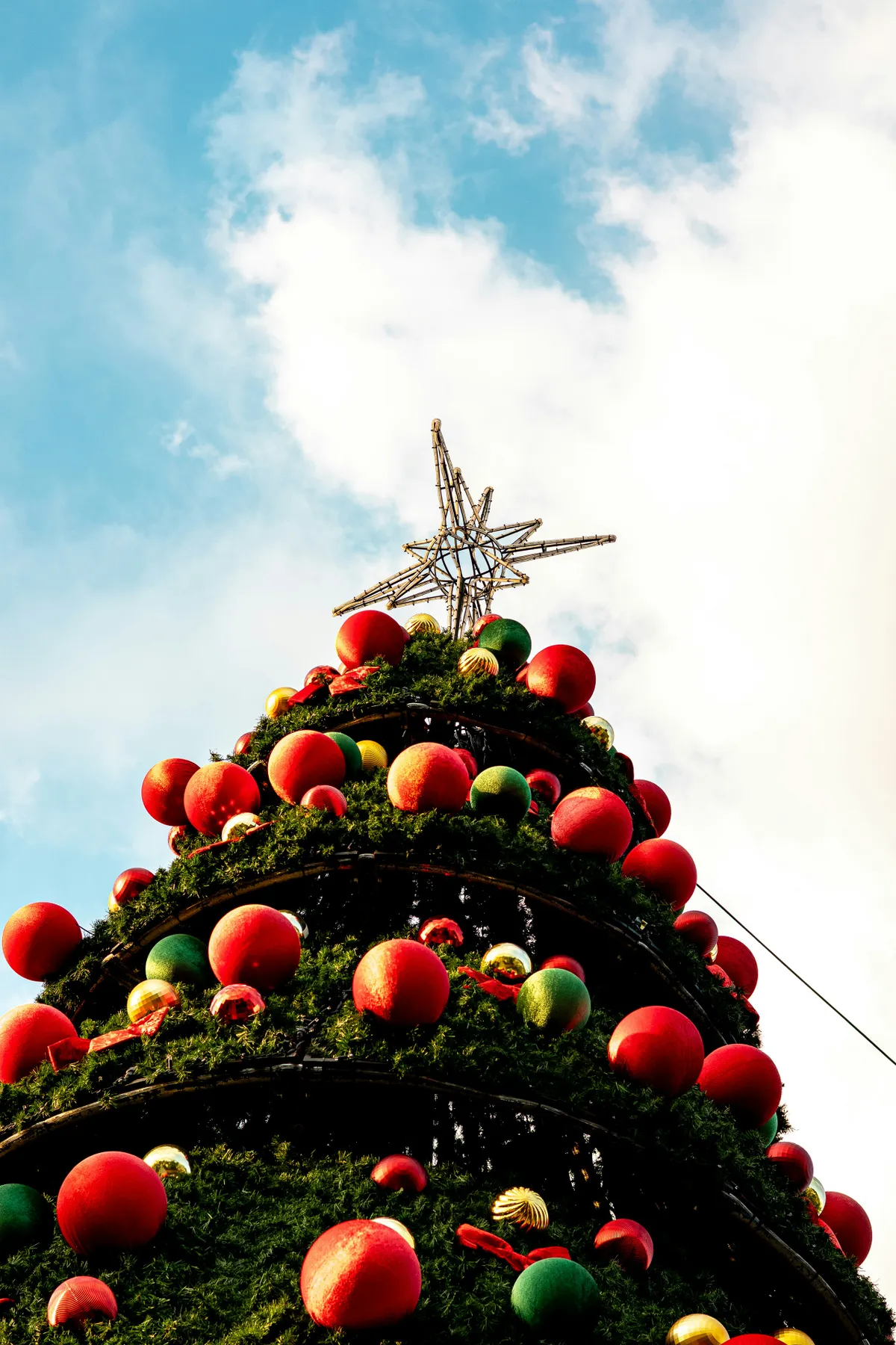 A large, decorated Christmas tree with red and green ornaments and a star on top, set against a bright blue sky with white clouds.