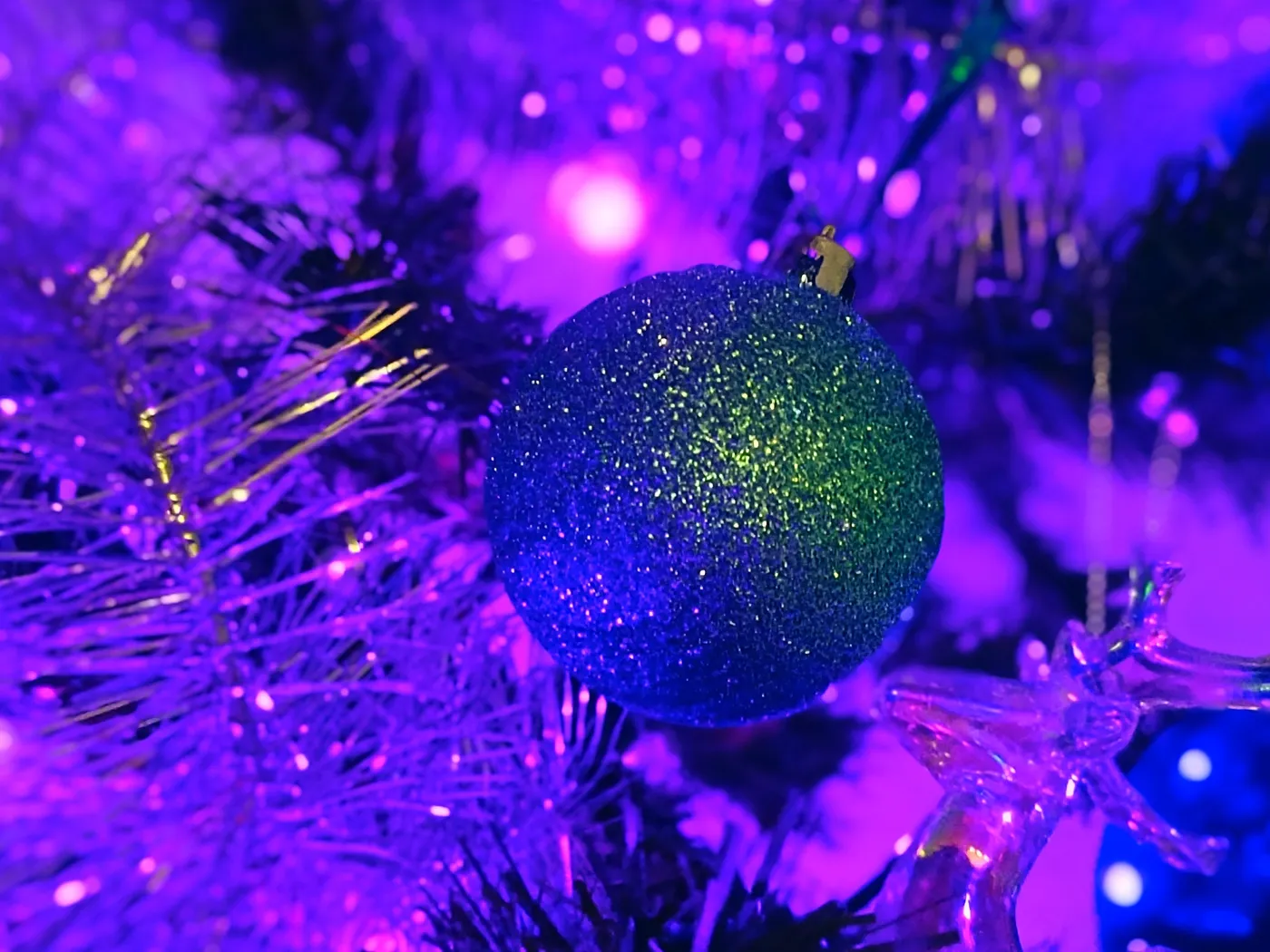A sparkling blue-green ornament hanging on a decorated Christmas tree with vibrant purple lights.