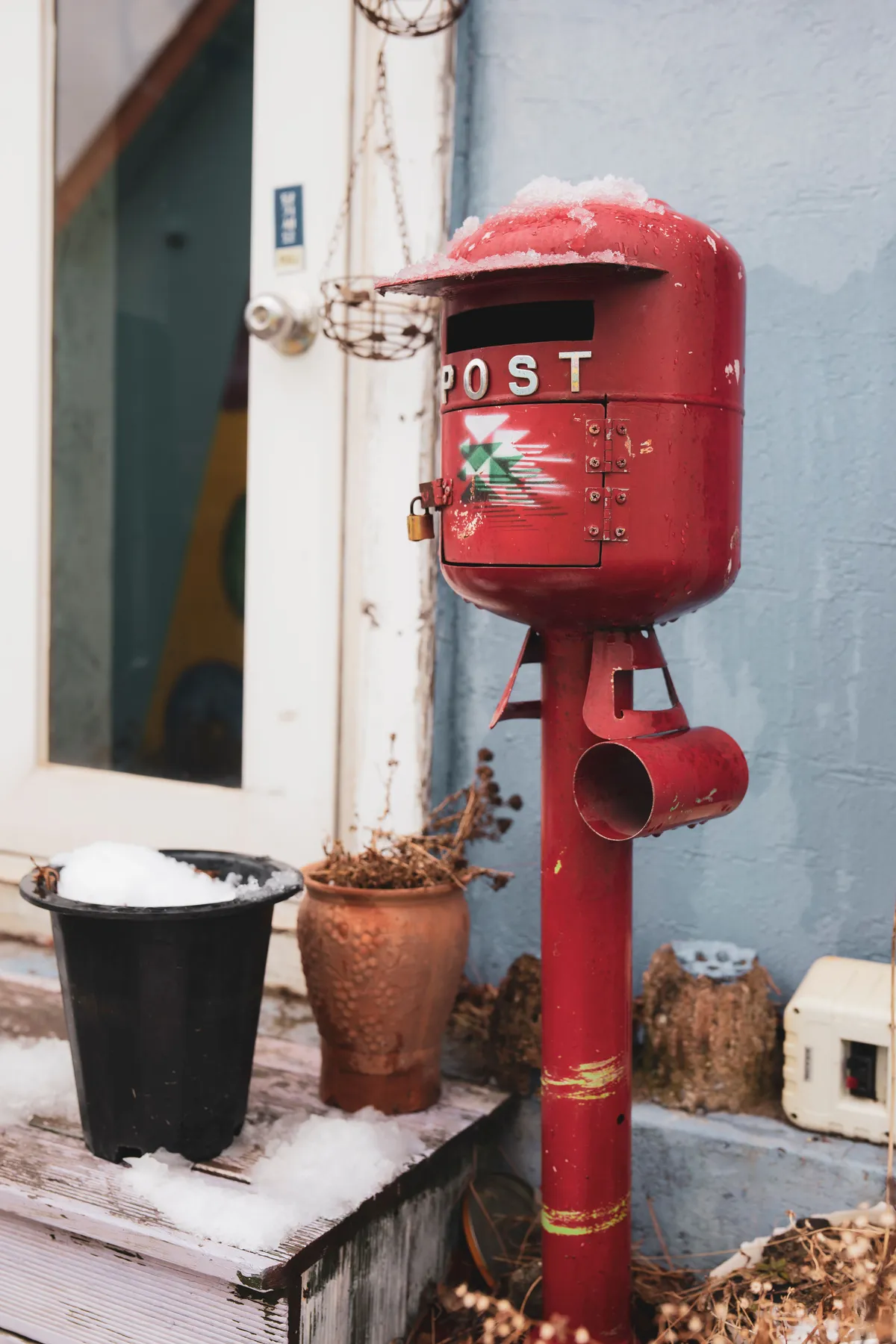 A vintage red post mailbox stands covered in snow beside potted plants outside a blue wall.