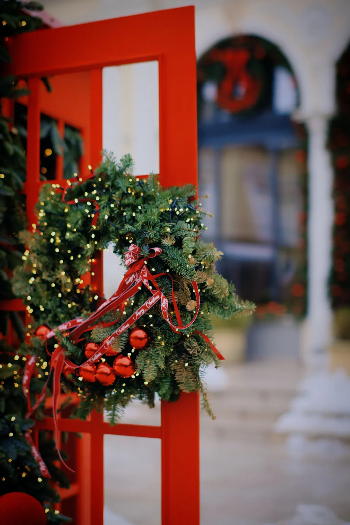 Festive Christmas wreath with red ornaments and ribbons on a red door frame, symbolizing holiday cheer.