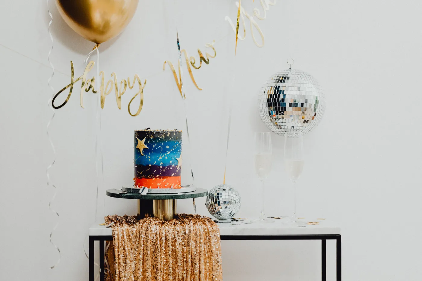 Glamorous New Year's party setup featuring a cake, balloons, confetti, and disco balls.