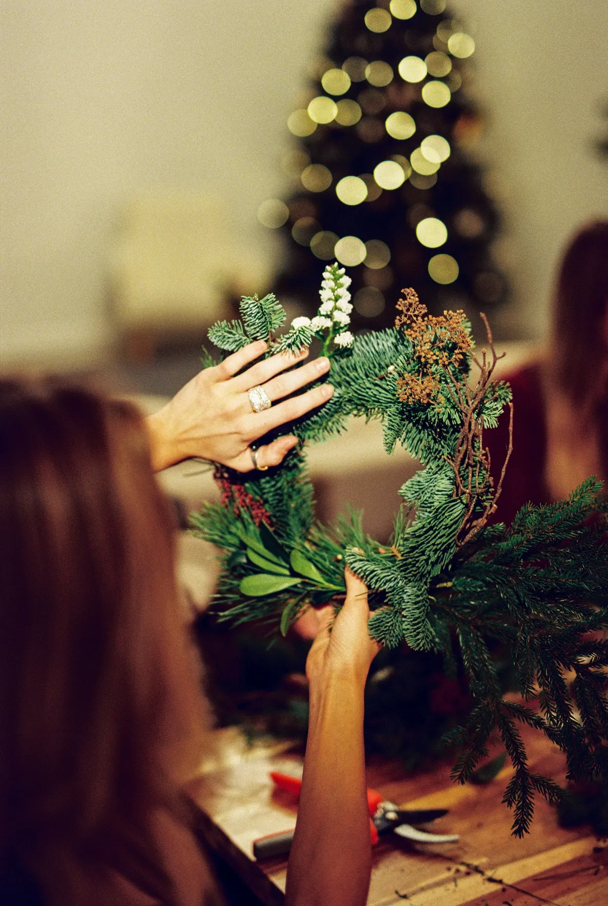 Hands creating a holiday wreath with a Christmas tree in the background, evoking a festive atmosphere.