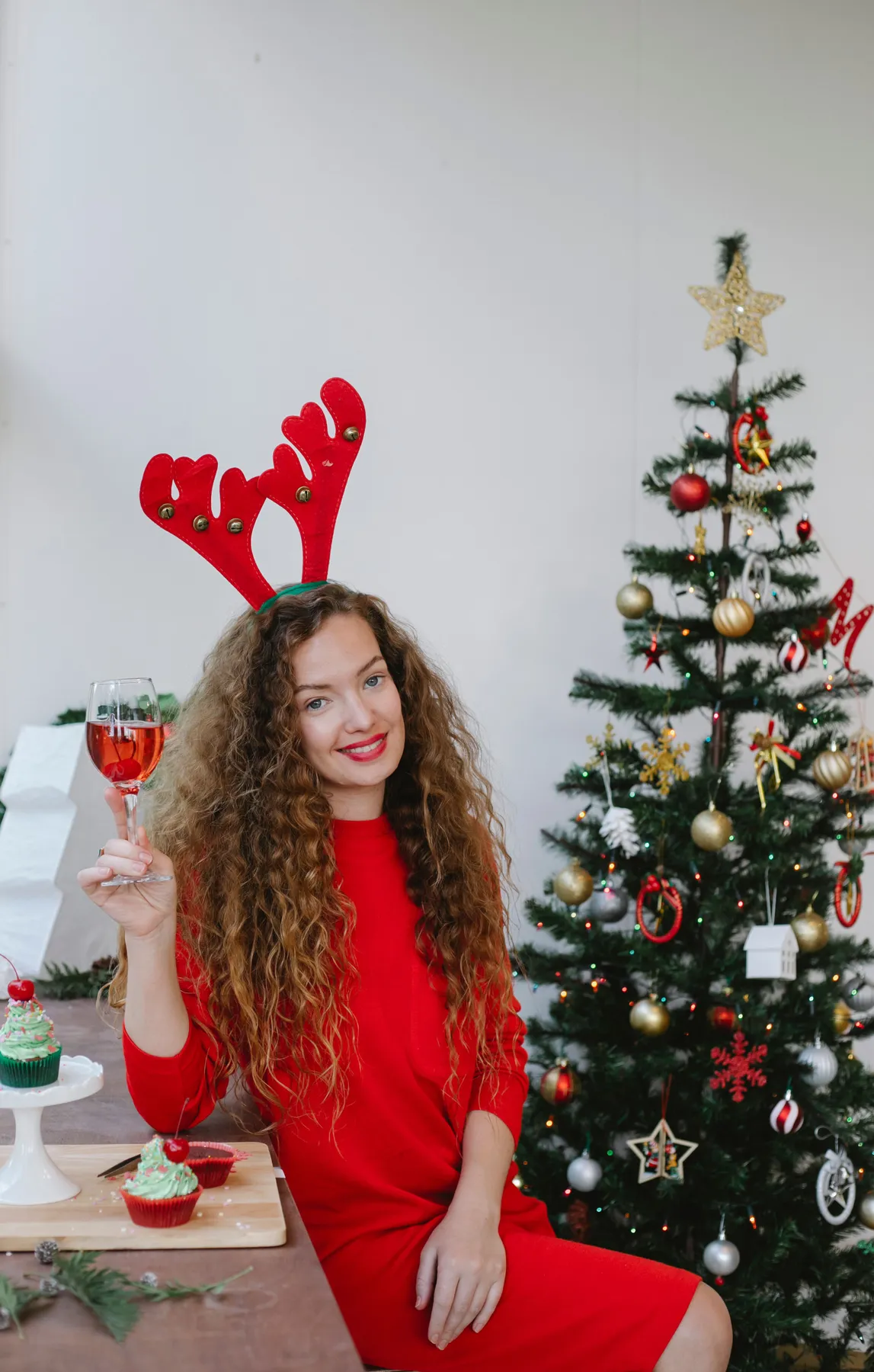 Happy woman with hairstyle in stylish clothes sitting with glass of champagne and celebrating Christmas in cozy room with festive decorations