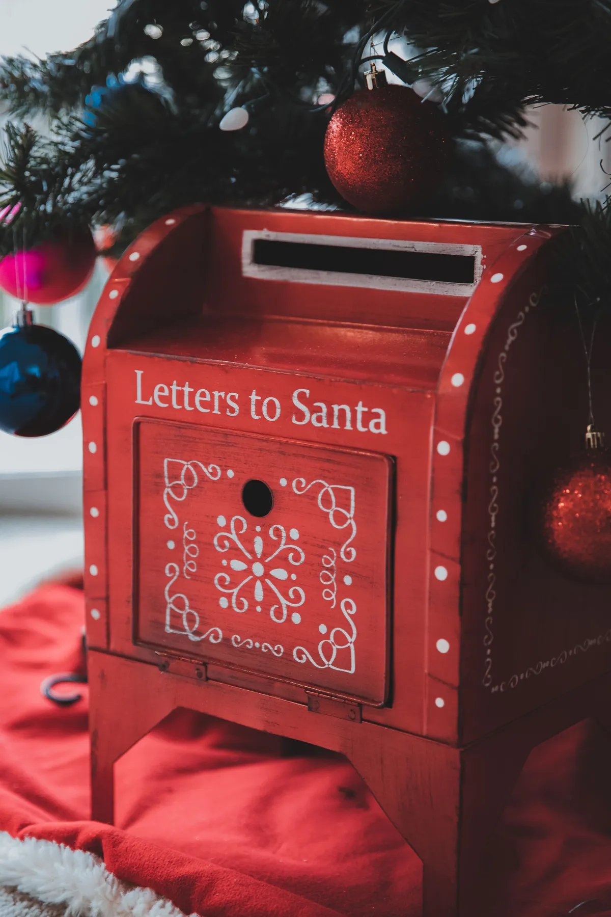 Red Christmas holiday mailbox for letters to Santa placed on rug under decorated Christmas tree branch with baubles in room