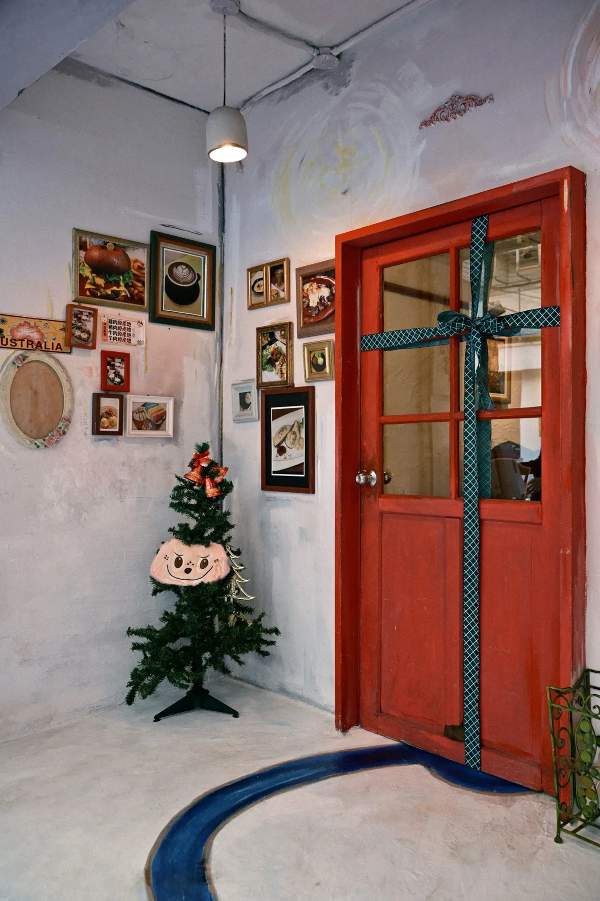 Red door wrapped in ribbon, small Christmas tree with decorations beside eclectic wall art indoors.