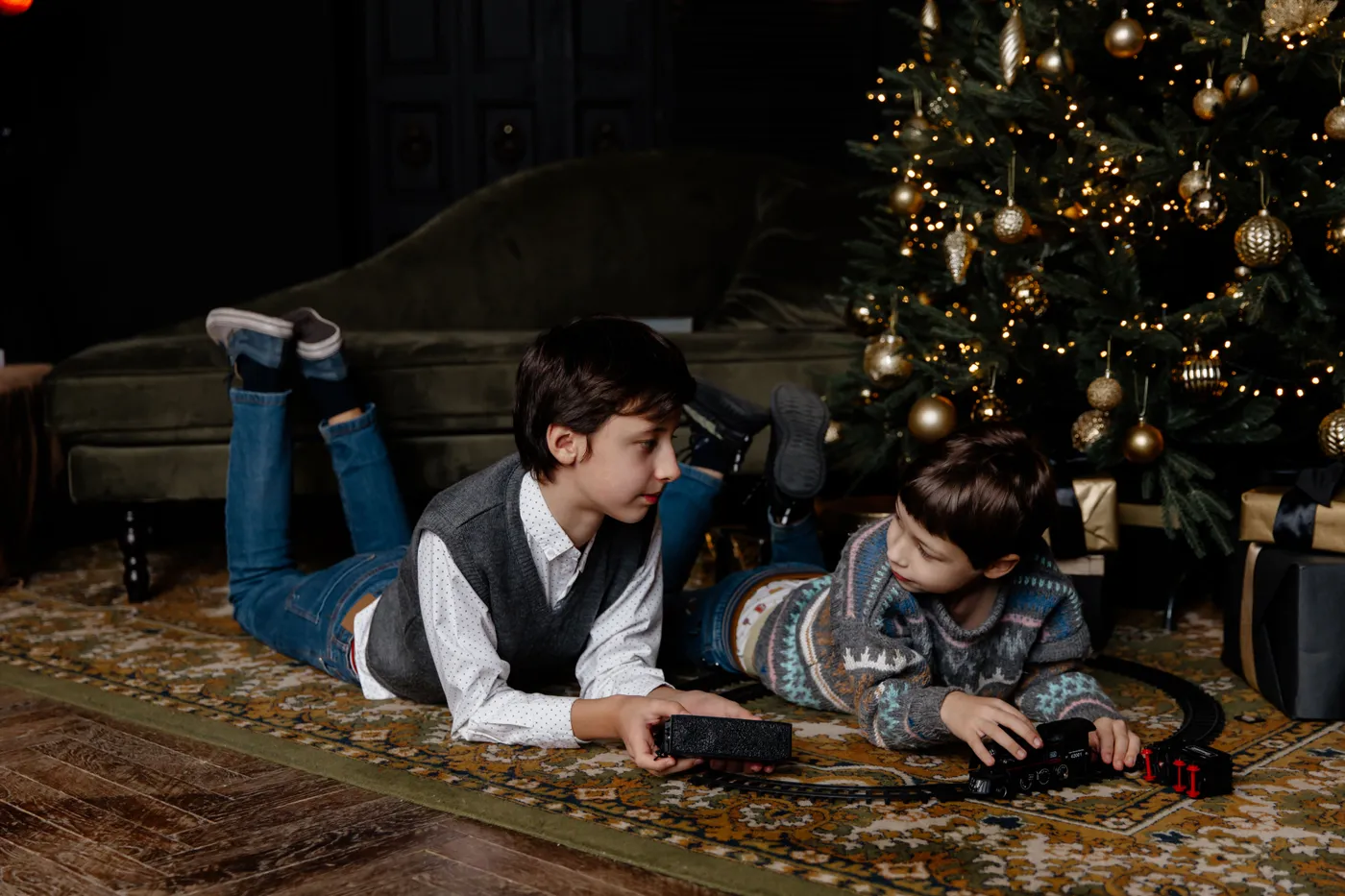 Two young brothers lying on a rug and playing with toys next to a Christmas tree indoors.